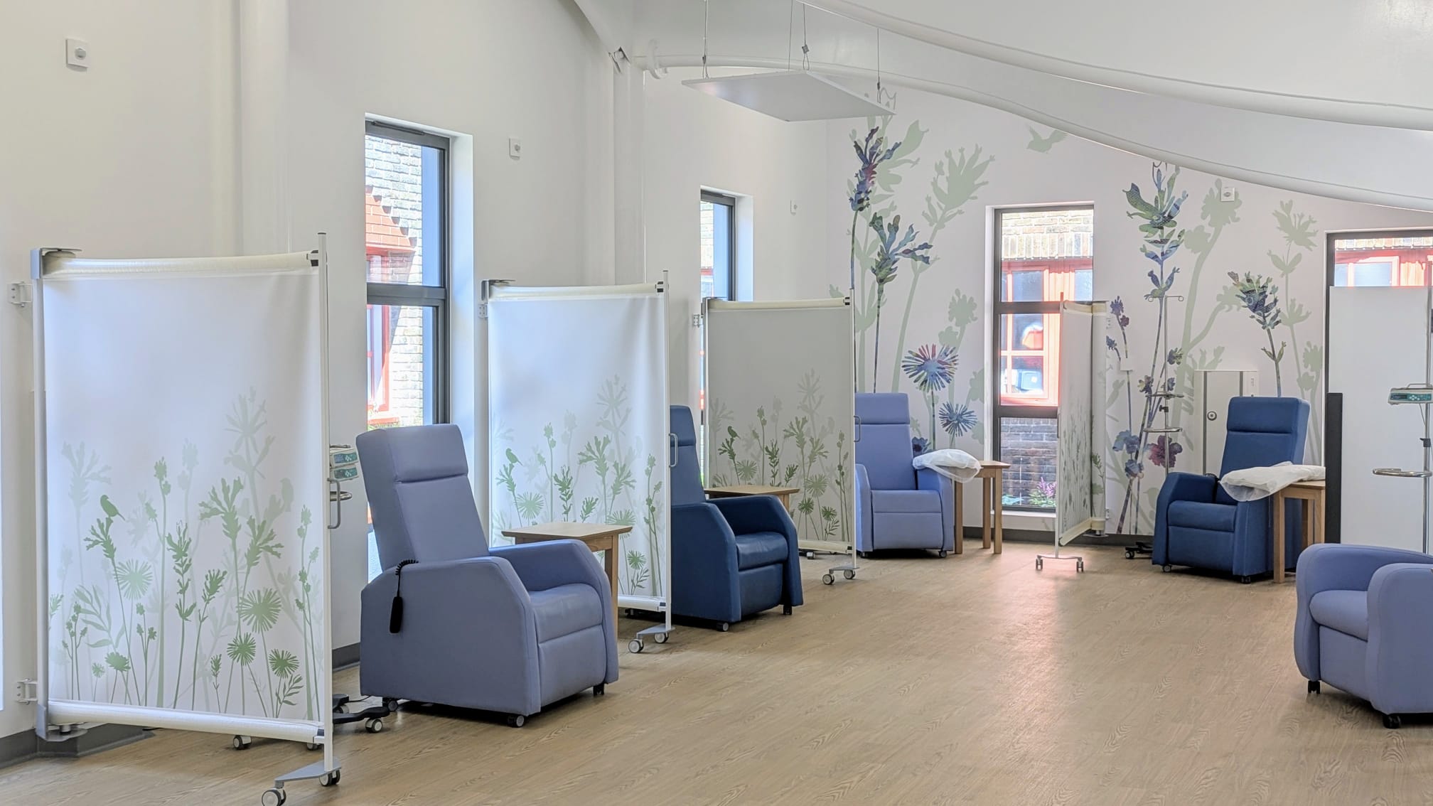 Interior of a bright and modern hospital room with blue chairs, white privacy screens decorated with green plant motifs, and large windows allowing natural light.