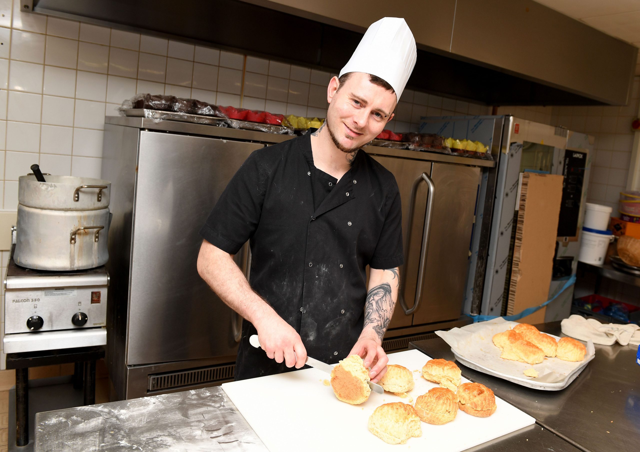 A chef standing in the kitchen, smiling at the camera. He is wearing a chef's hat and slicing bread.