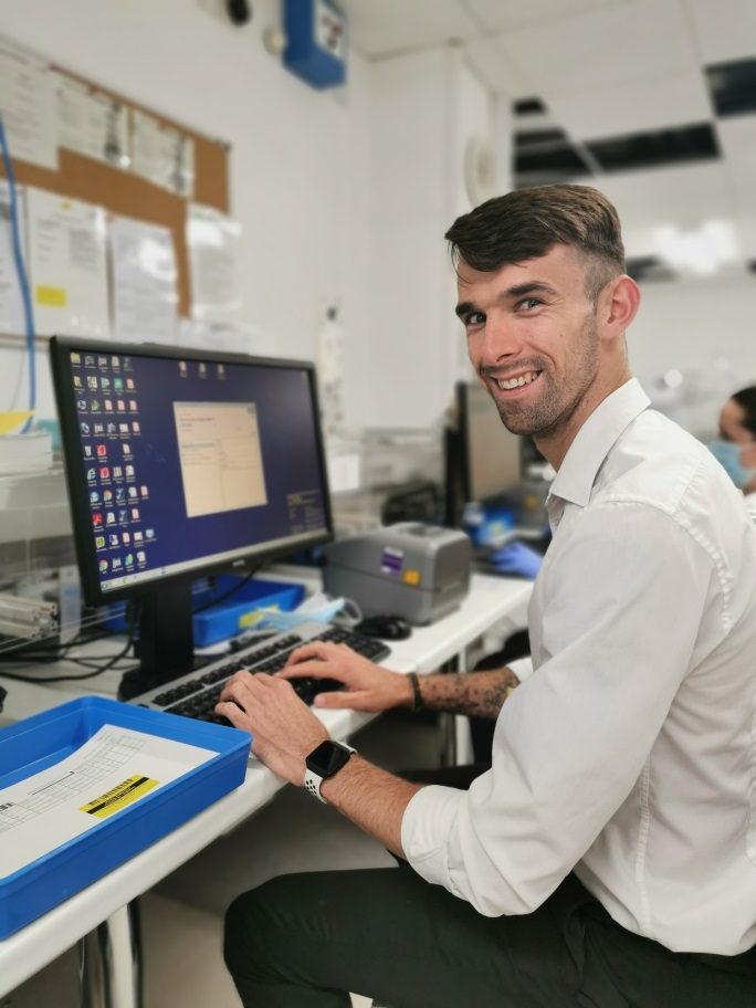A person in a white shirt sitting at a computer. He is turned to the side and smiling at the camera.