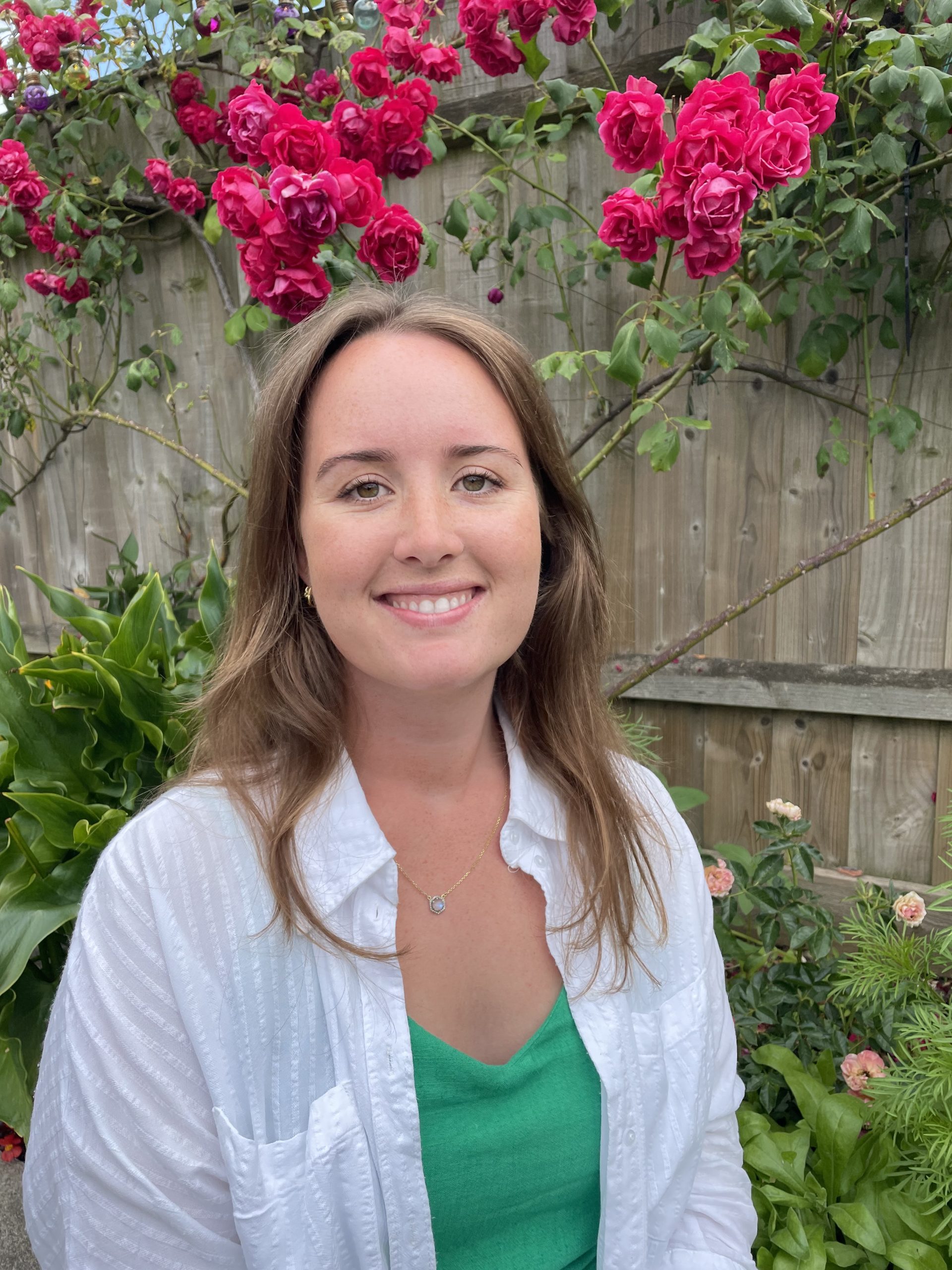 A woman smiling at the camera. She is sitting in a garden with pink flowers behind her.