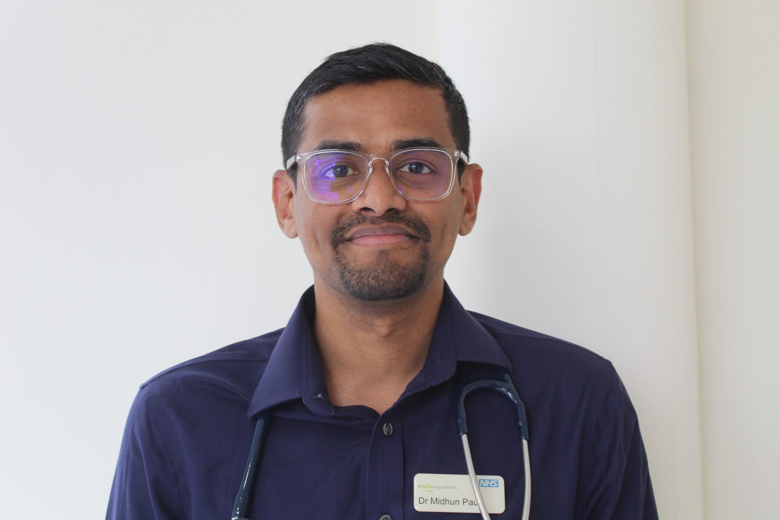 A person standing, smiling at the camera. They are wearing a shirt and ID badge.