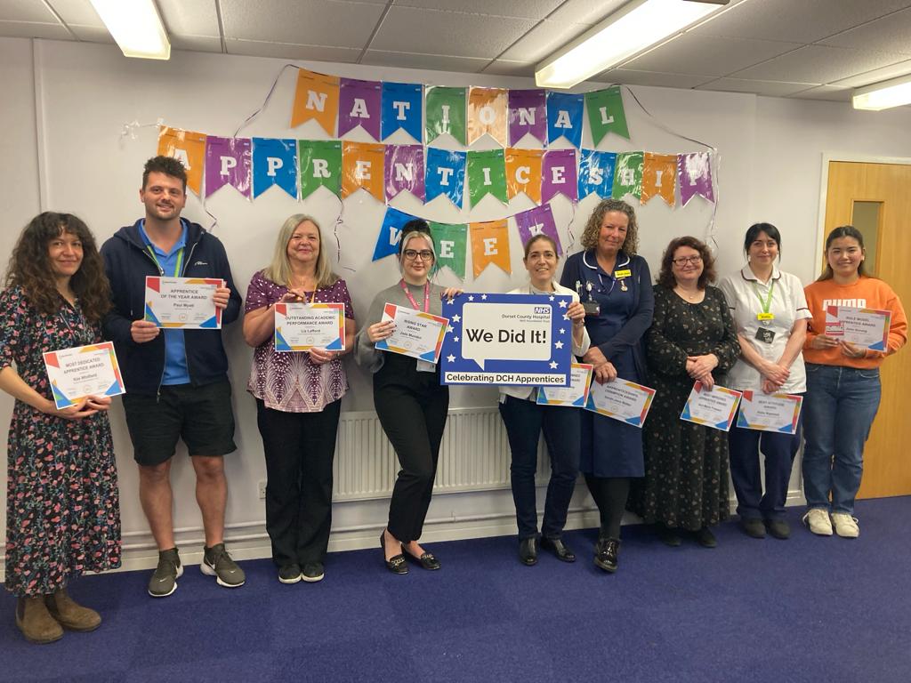 A group of staff pictured holding certificates. They are standing in front of a banner that reads 'Apprenticeship Week'.