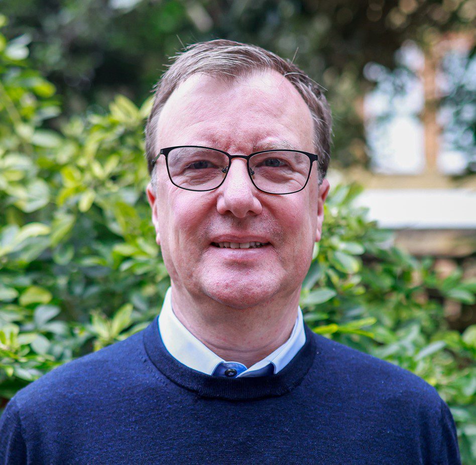 A man standing outside smiling at the camera. He is wearing a blue shirt and jumper and glasses.