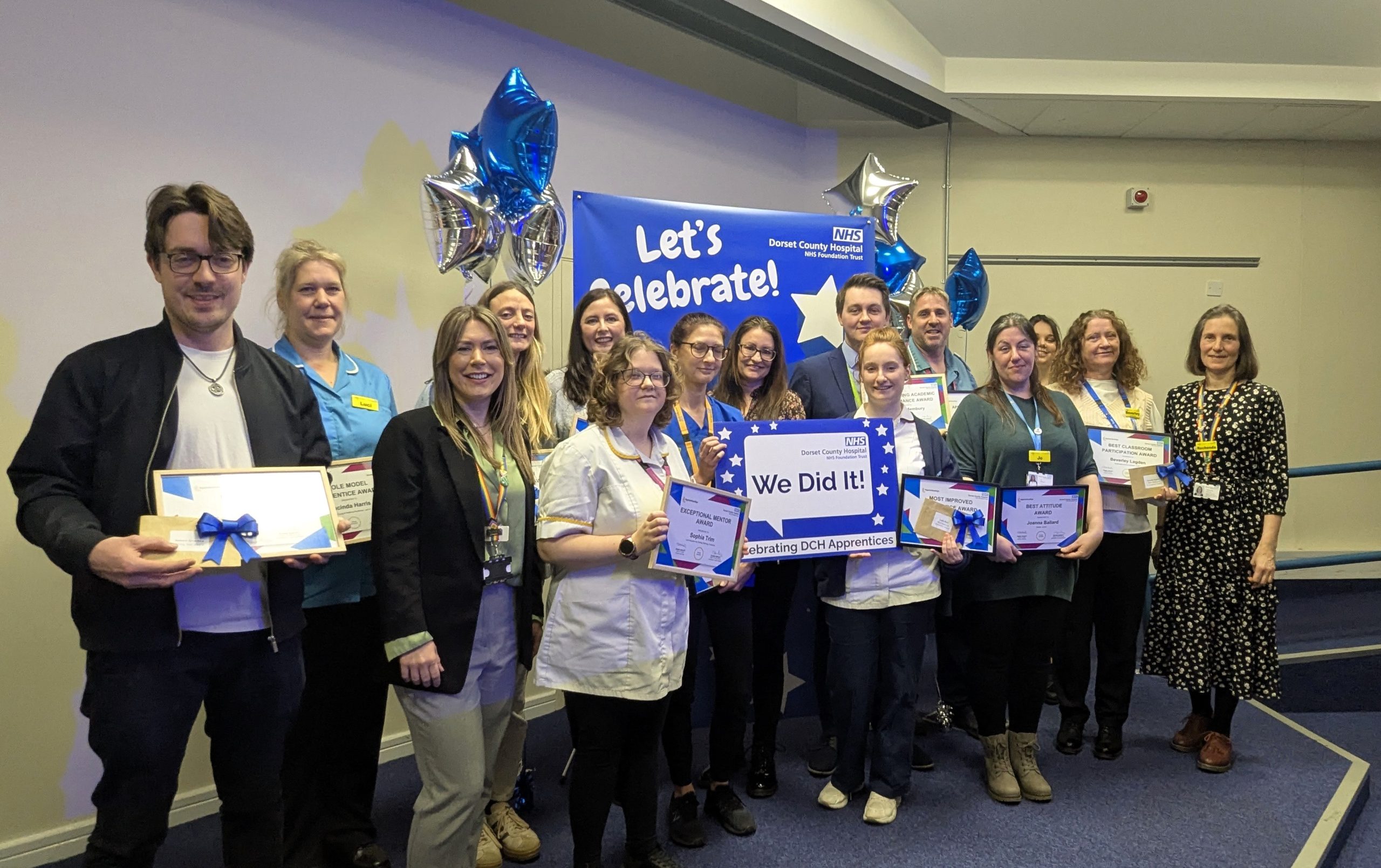 A group of healthcare staff standing together holding certificates. There is a banner in the background that reads: "Let's Celebrate" as well as blue and silver star balloons.