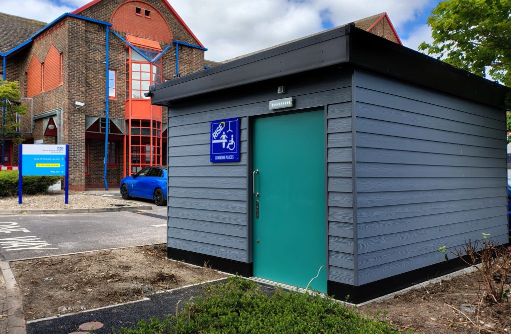 Outdoor view of a grey Changes Places accessible toilet facility. There is a hospital in the background.