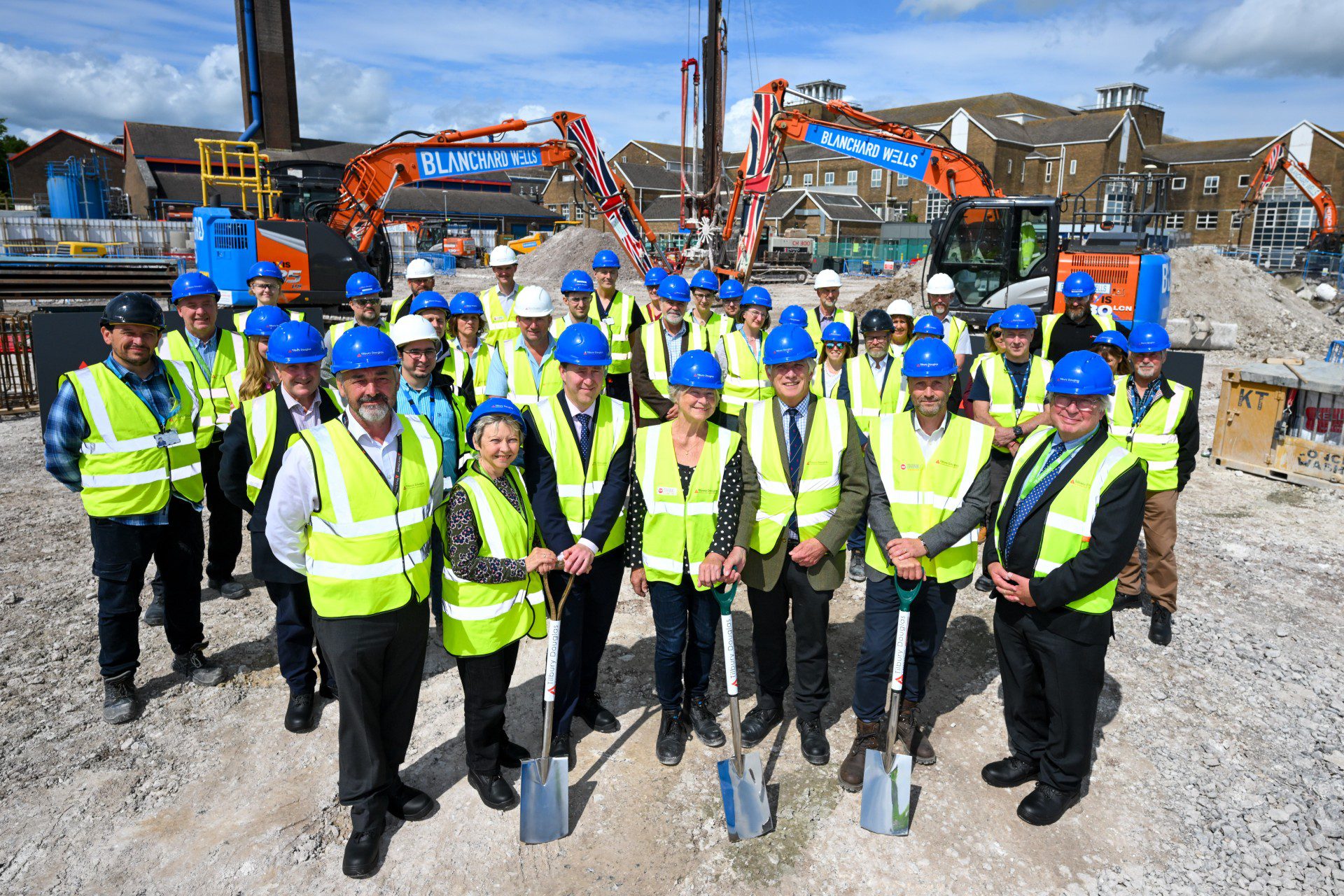 Group of people in hard hats and reflective vests, holding shovels at a construction site with heavy machinery and buildings in the background.