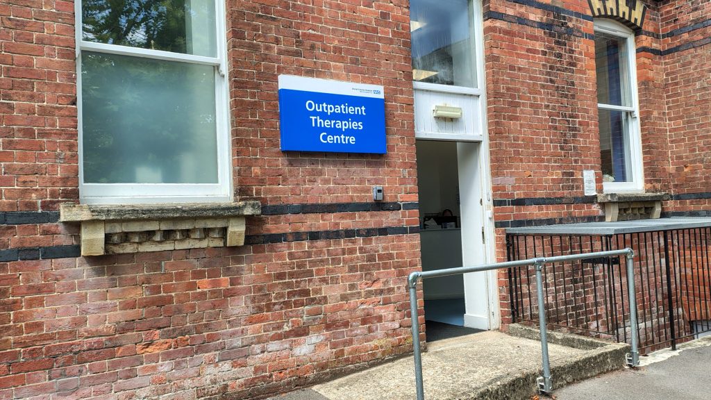 A sign for 'Outpatient Therapies Centre' mounted on a brick building with a ramp leading to a doorway.