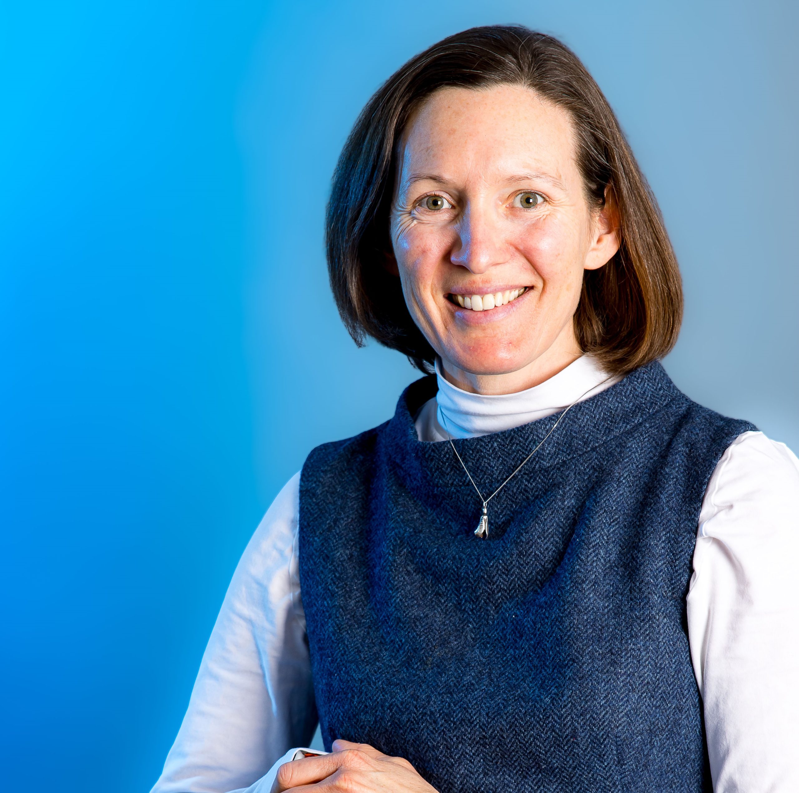 A woman in a professional dress, smiling at the camera. She is leaning on a chair.