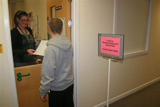 A person in a grey jumper at a reception desk. A sign reads 'Clinical Neurophysiology Reception, please wait here'.