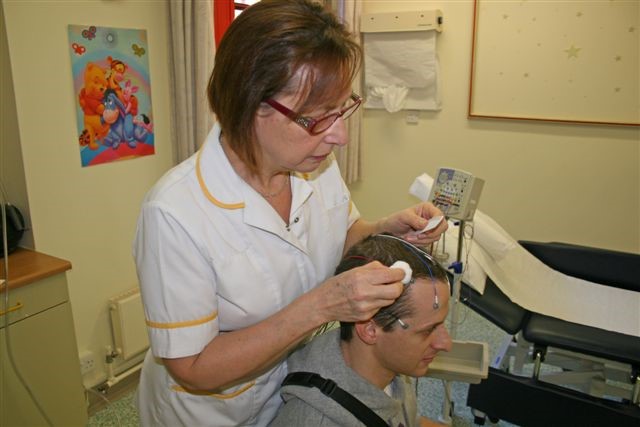 A healthcare worker is helping to remove wires from the top of a patient head, using a cotton bud and liquid.