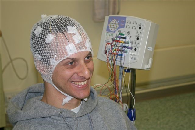 A person with wires connected to their head for a test in a hospital. A machine is in the background.