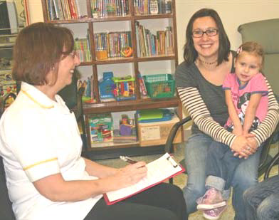 A young girl sitting on her mother's knee. A healthcare worker is sitting opposite with a clipboard.