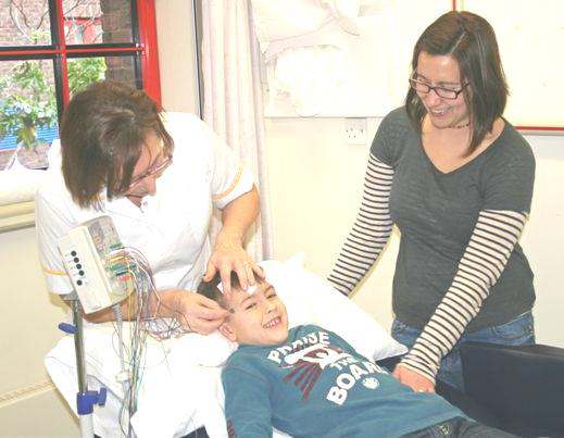 A young boy is lying on a hospital bed. His mother is standing next to him smiling while a healthcare worker is attaching wires to the top of his head.
