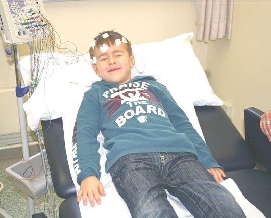 A child is lying on a hospital bed. He has wires stuck to the top of his head and has his eyes closed.