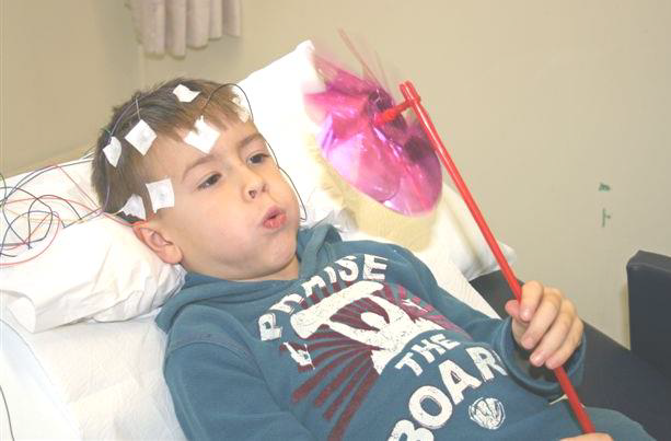 A child is lying in a hospital bed blowing at a spinning toy.