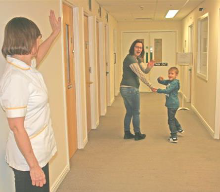 A healthcare worker waves goodbye to a child and her mother in a hospital corridor.