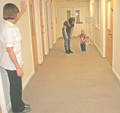 A healthcare worker waves goodbye to a child and her mother in a hospital corridor.