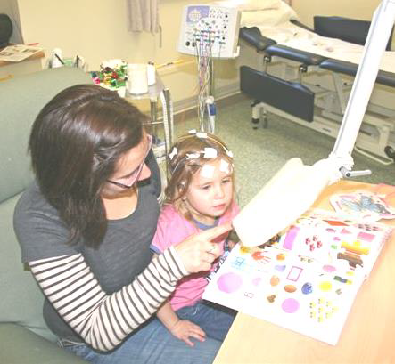 A child is sitting on her mother's knee in a clinical setting. A light is flashing in front of her.