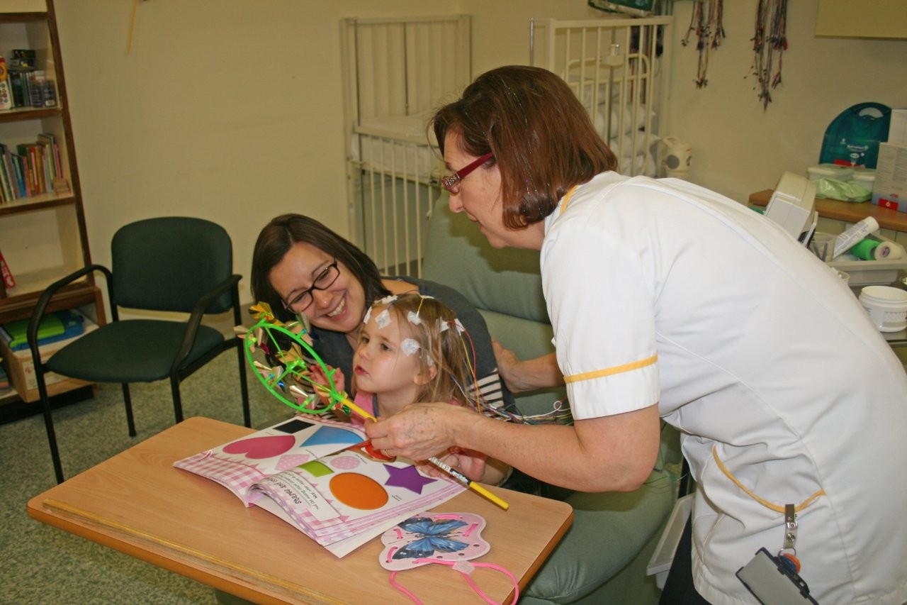 A child is sitting on her mother's knee in a medical setting. She is blowing a spinning toy.