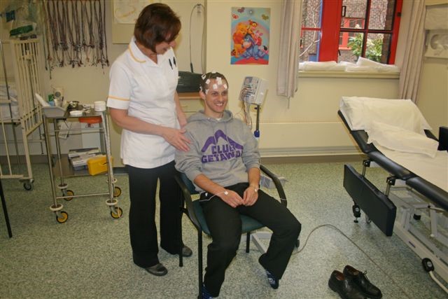 A patient sitting in a hospital room. They have wires on their head.