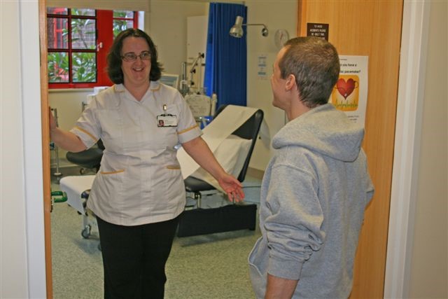 A healthcare worker smiling at a patient. They are holding the door open as if they are saying goodbye. Behind them is a hospital examination bed.