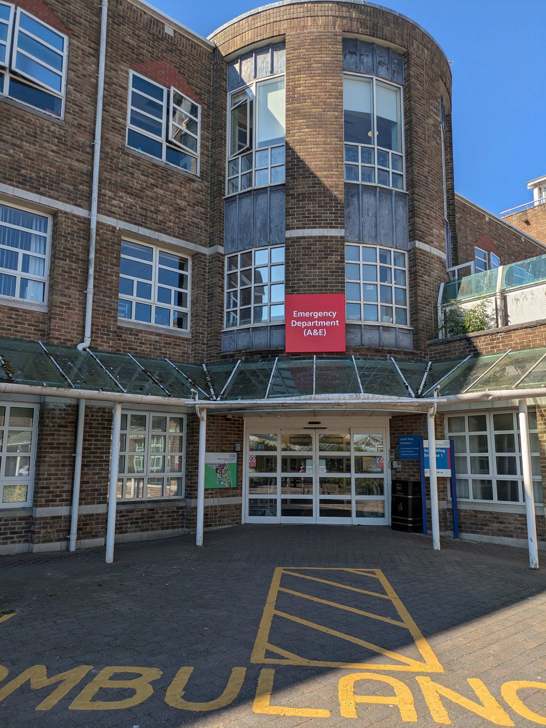 An exterior image of a building. A red sign that reads 'Emergency Department (A&E)' is written above the door. There is also yellow markings on the floor outside the building, indicating parking spaces for ambulances.
