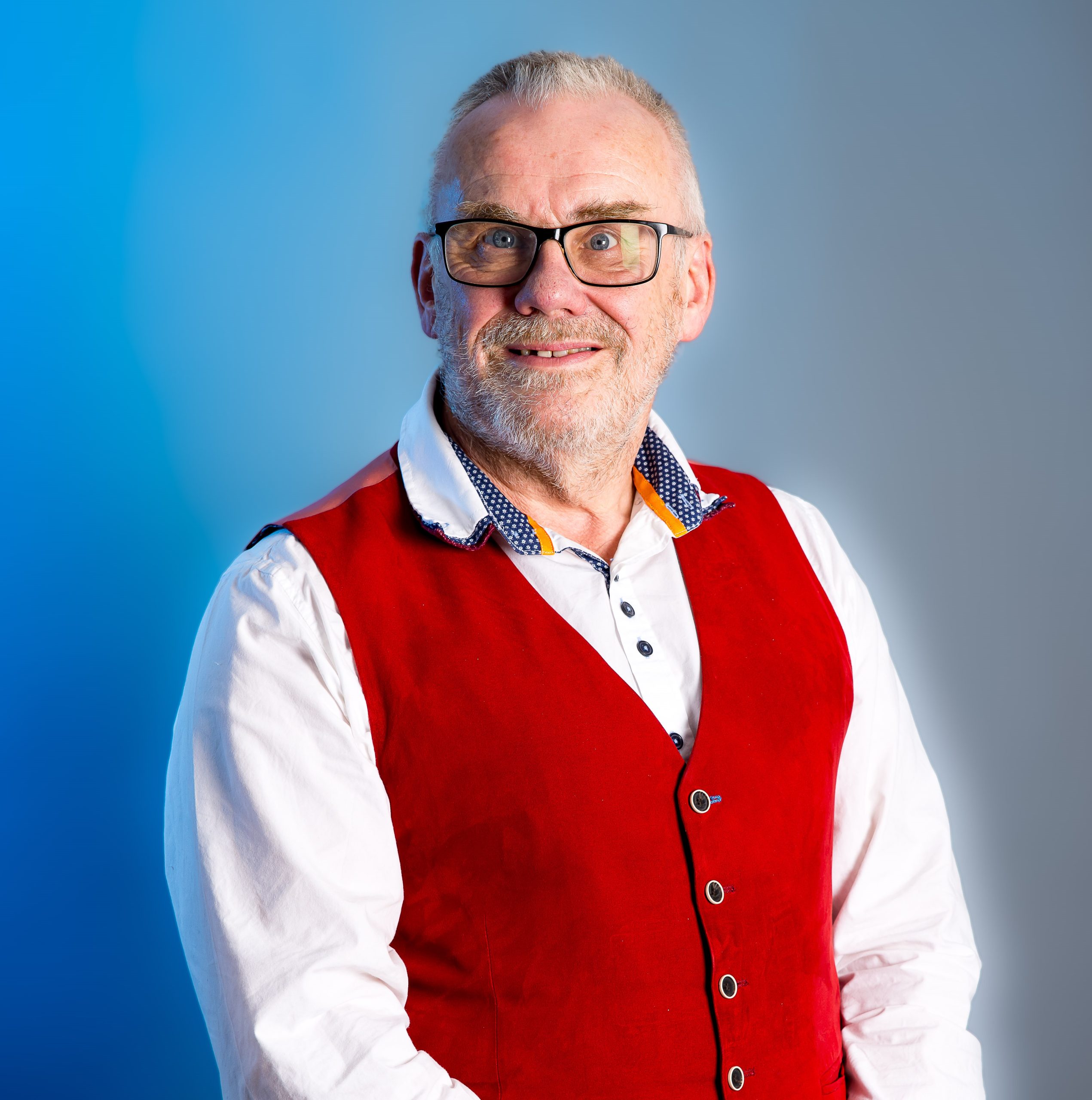 A man smiling at the camera. He is wearing a white shirt and red waistcoat.