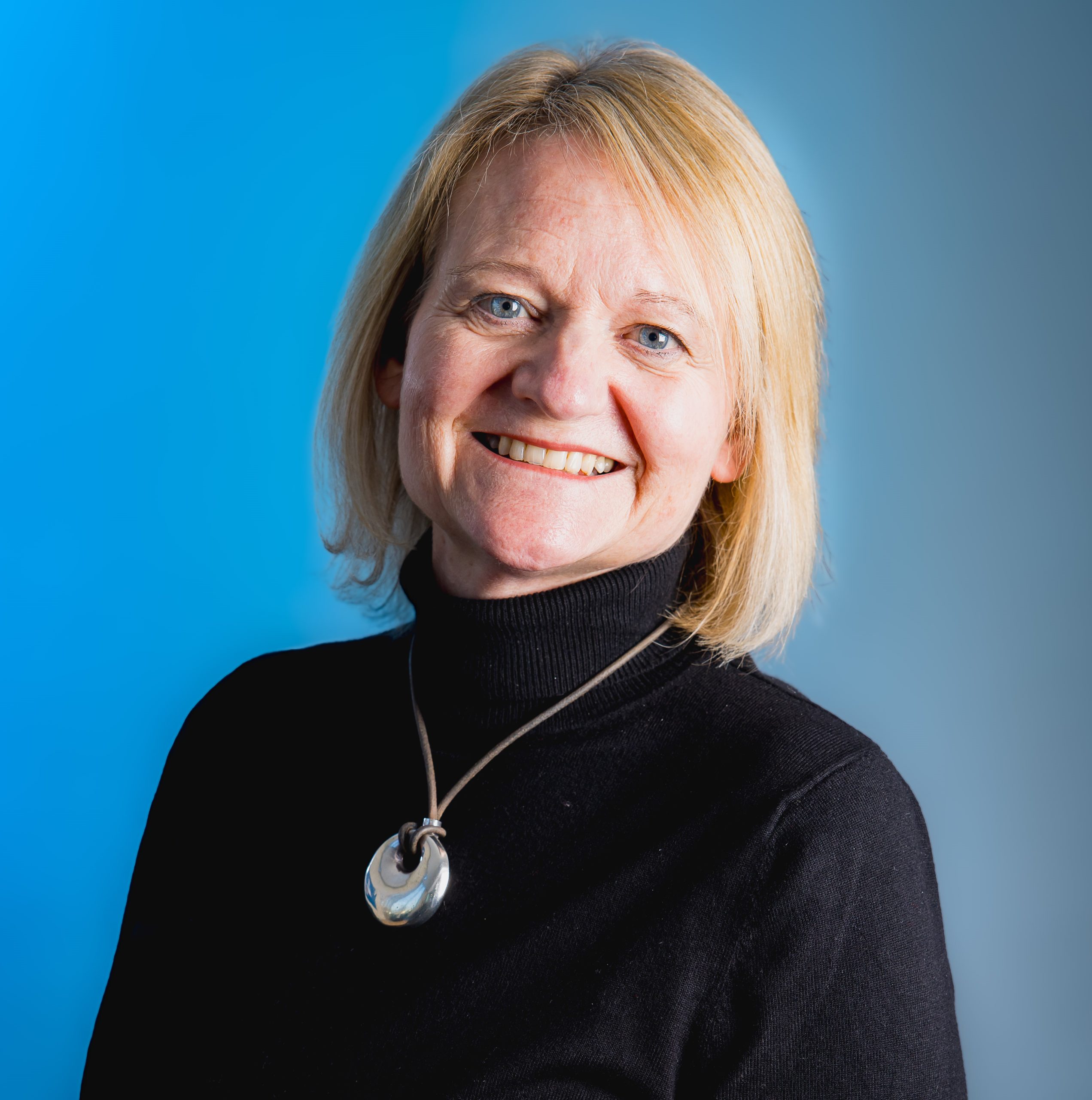 A person smiling at the camera. She is wearing a black jumper and a chunky necklace.