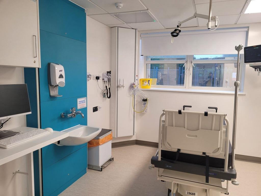A brightly lit room in a hospital. There is medical equipment in the room, as well as a bed, sink and computer.
