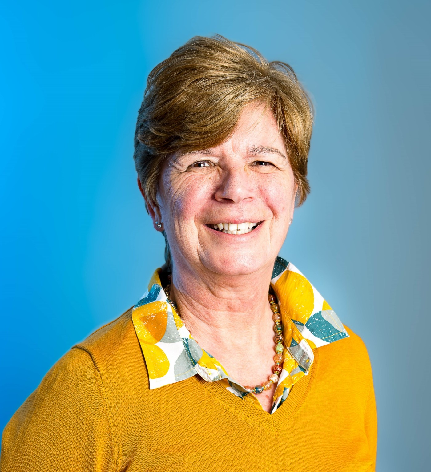 A woman smiling at the camera. She is wearing a yellow and blue shirt with a yellow jumper and a necklace.