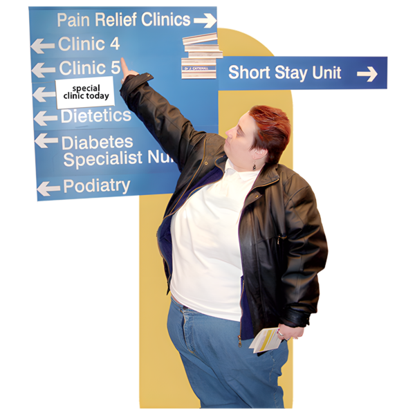A person pointing to a directional sign in a healthcare facility with options for Pain Relief Clinic, Diabetes Specialist Nurses, and Podiatry; other signs indicate Clinic 4 and Short Stay Unit directions.