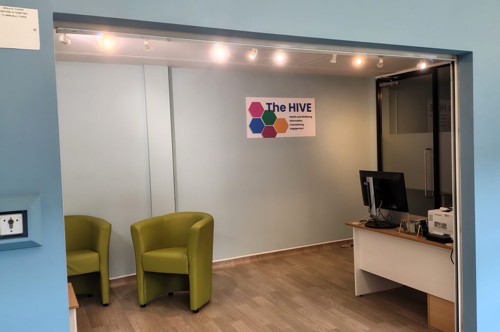 A modern office area featuring two green chairs, a desk with a computer, and a sign that reads 'The HIVE' on the wall.