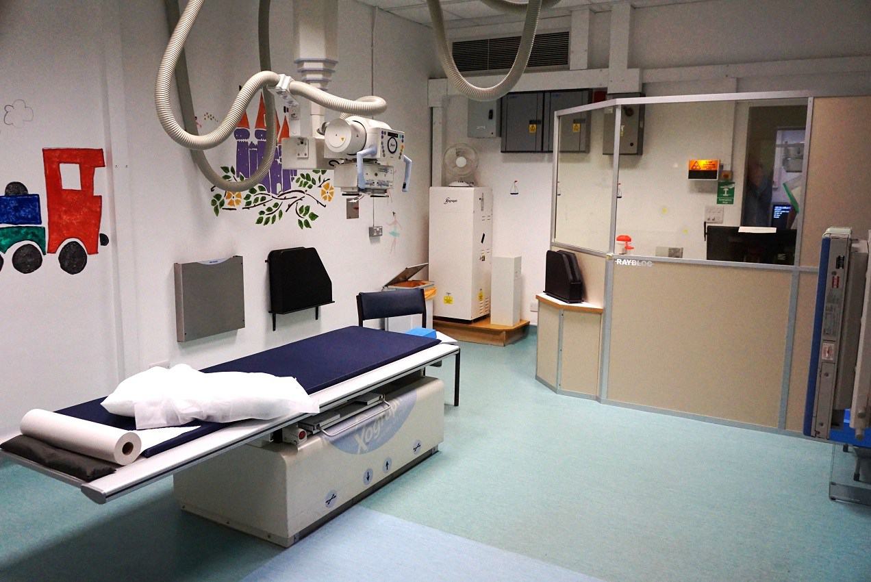 A brightly lit room in a hospital. Above a bed is a machine used for x-rays. There area colourful pictures drawn on the wall.