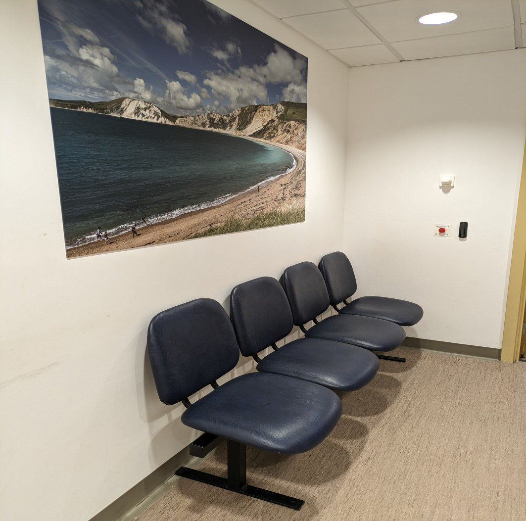 Four blue seats in a corridor. Above the seats is a scenic photo.