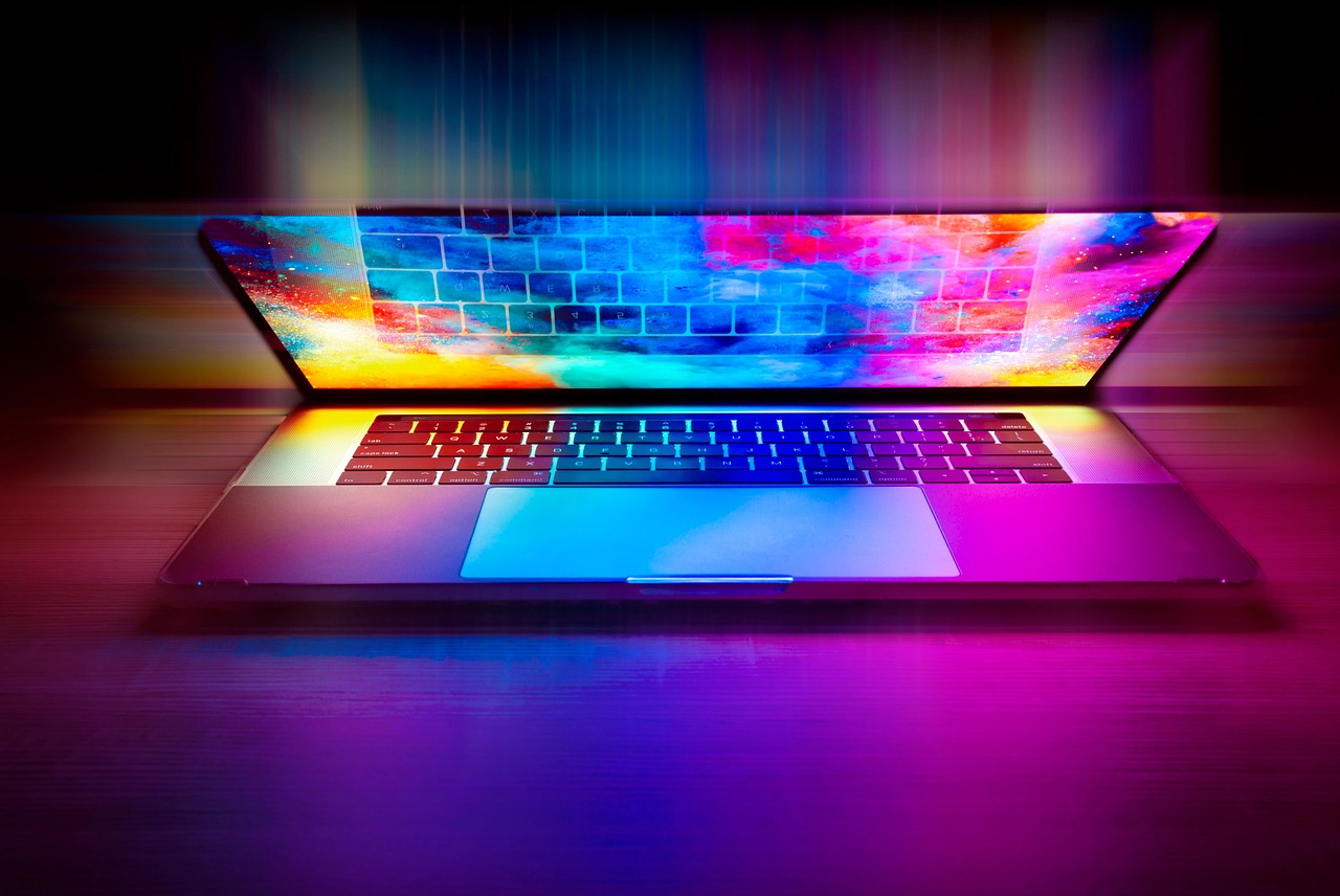 An open laptop displaying vibrant, colourful abstract wallpaper on screen with a glowing keyboard setting on a wooden surface, illuminated by neon lights.