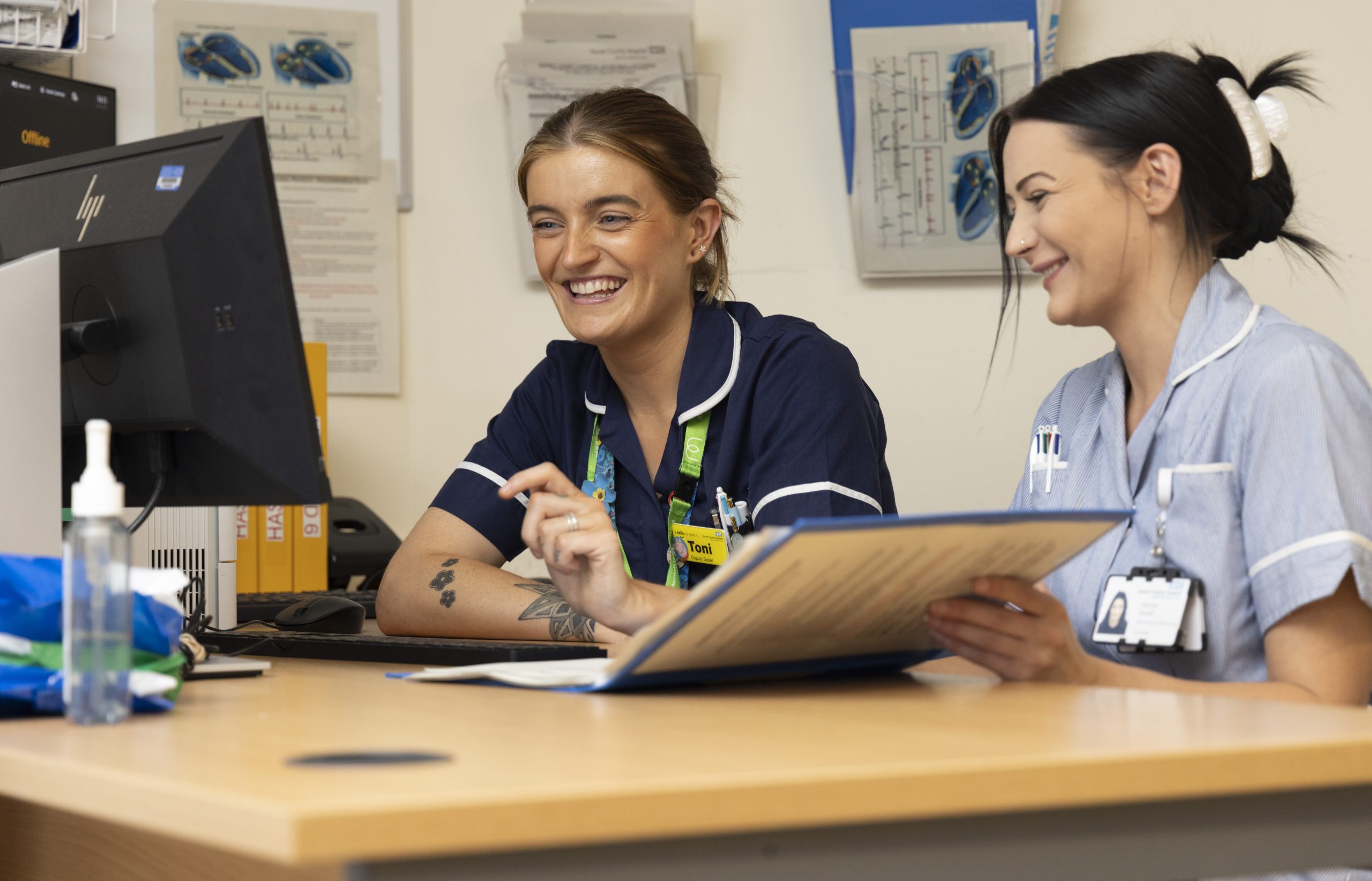 Two nurses sitting at a desk smiling. One is opening a folder.