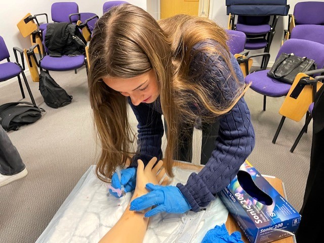 A person in a classroom setting, wearing gloves and practicing a medical procedure on a training arm.