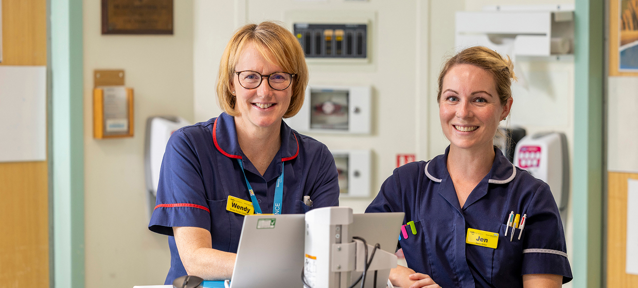 Two nurses standing at a nurses station on a ward, smiling at the camera.