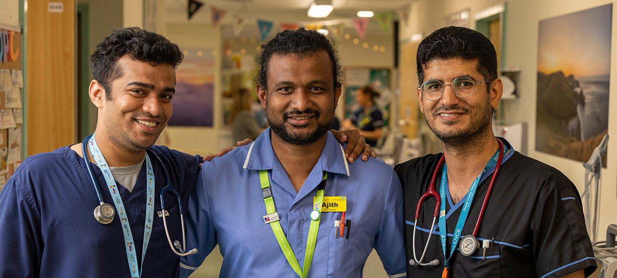 Three healthcare works standing together, smiling at the camera. Two of them have stethoscopes round their necks. They are standing in the ward of a hospital.