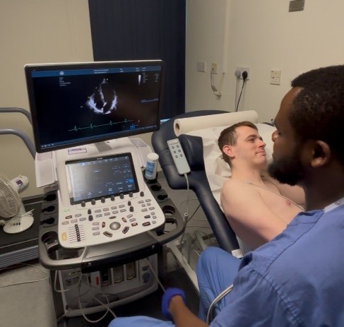 A person lying in a hospital bed with a healthcare professional sitting next to them. The patient is undergoing some cardiac tests.