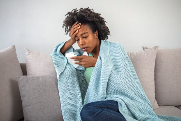 A person wrapped in a blue blanket sitting on a couch, looking unwell and holding a tissue.
