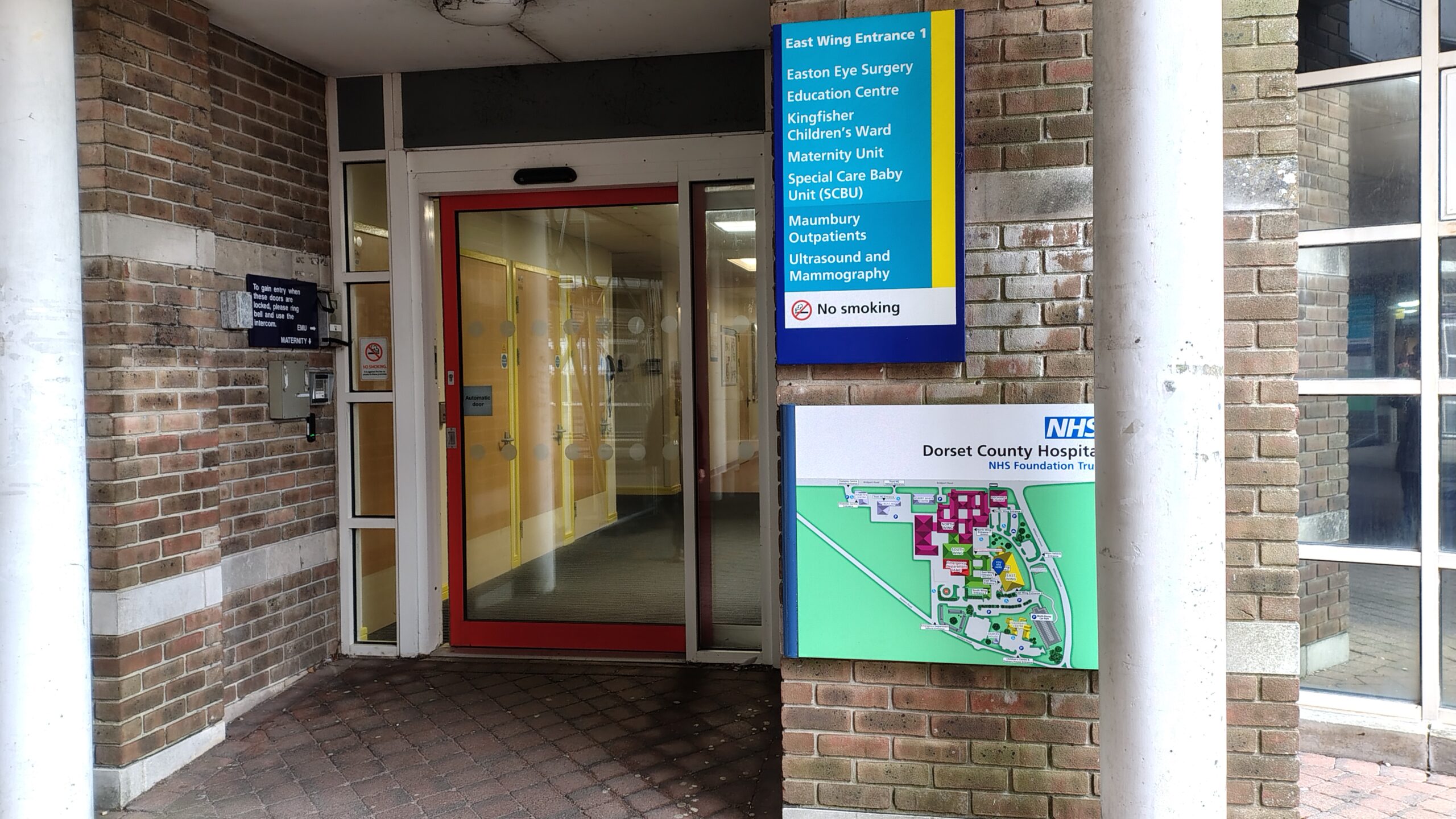 An entrance of a hospital. Outside the door is a hospital a map and a sign titled 'East Wing Entrance 1'.