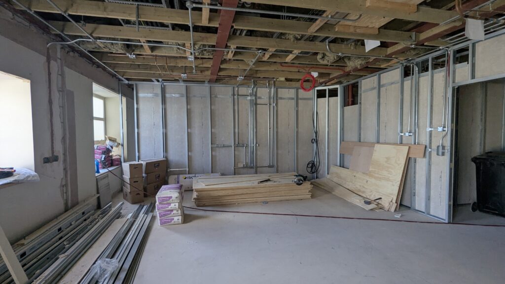 An interior of a room under construction with exposed wooden beams, insulated walls, and construction materials such as wooden planks and boxes on the floor.
