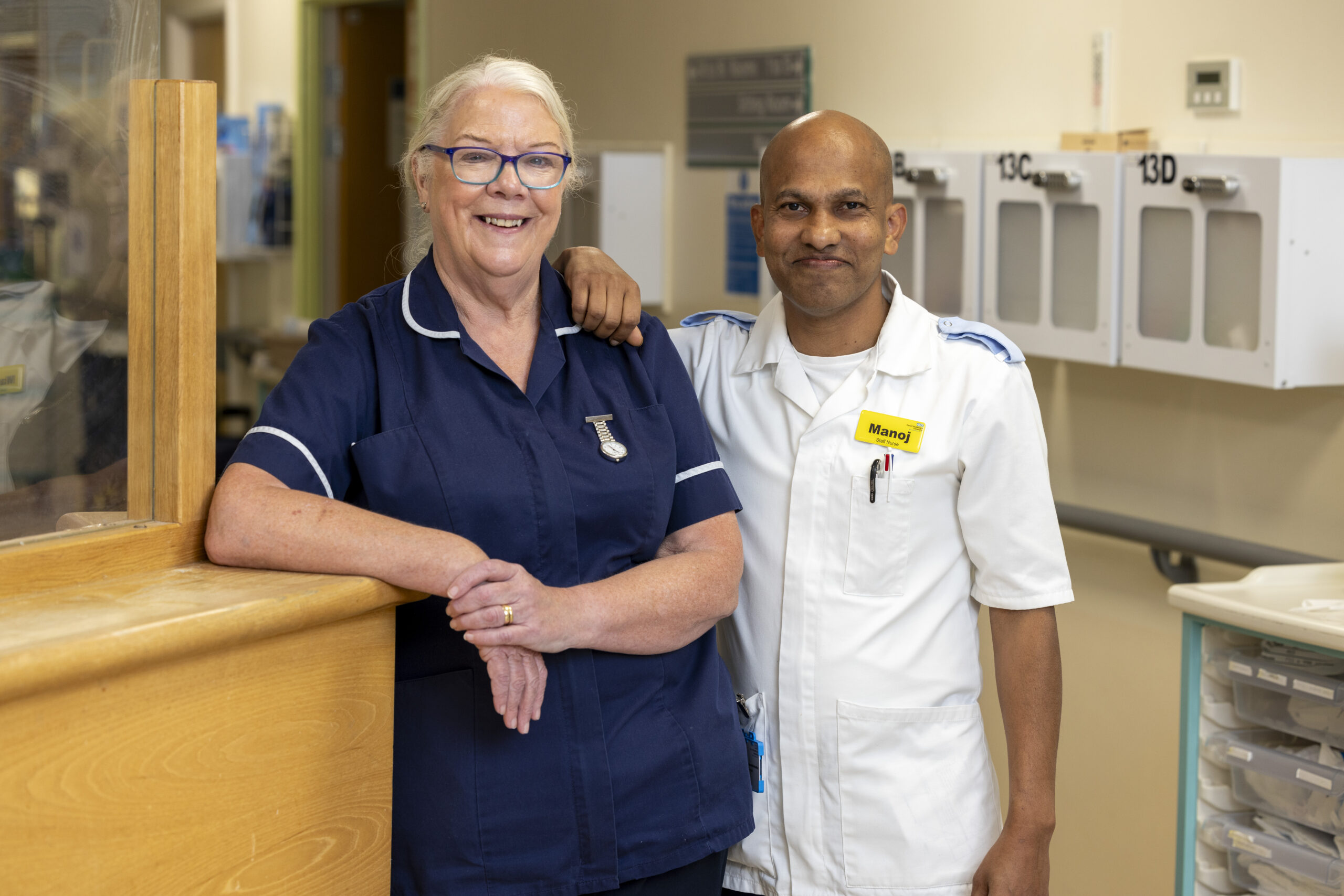 Two heatlhcare workers standing together in a hospital corridor, they are both smiling at the camera.