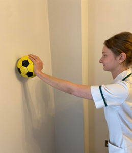A healthcare professional holding a yellow ball against a wall with one hand.