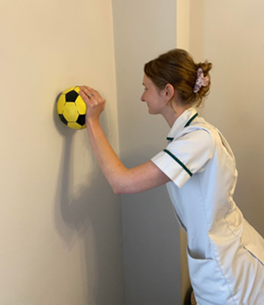 A healthcare professional holding a yellow ball against a wall with one hand.