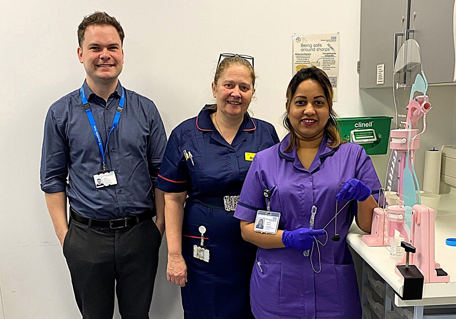Three healthcare professionals are standing in a medical procedure room. They are wearing uniforms with identification badges. Behind them is a cart with medical equipment.