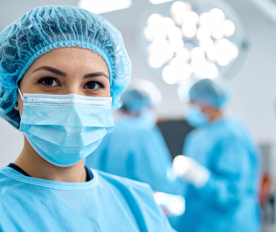 A medical personnel in surgical attire, including masks and hair covers, are in an operating room with bright surgical lights. One person is in the foreground, while others are blurred in the background during a procedure.