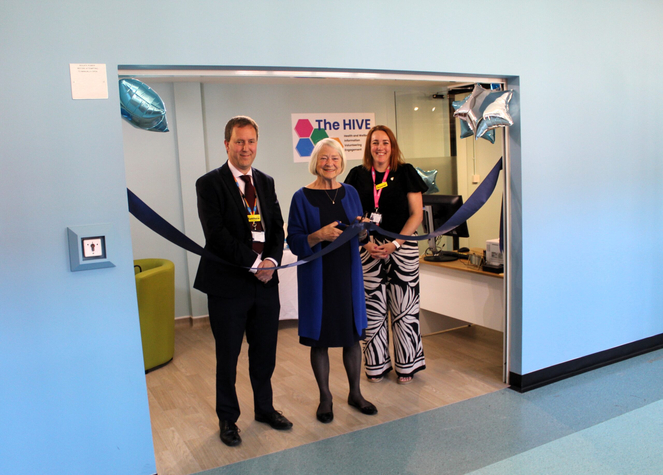 The people standing in the doorway of a new office space. One person is holding a pair of scissors and is about to cut a ribbon.