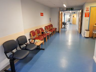A corridor in a hospital with chairs lined up against the wall. There is a small table with magazines and leaflets on it.
